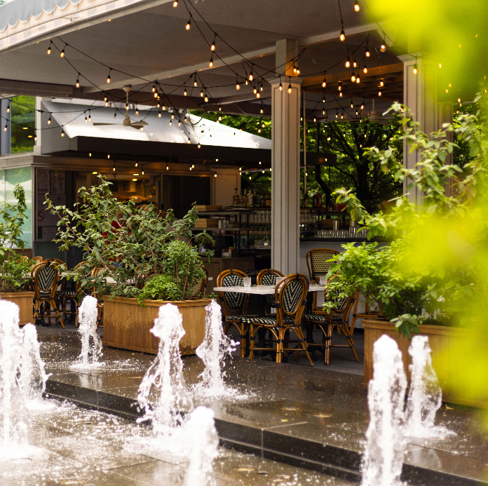 An inviting outdoor restaurant patio surrounded by lush greenery and large potted plants, with rattan bistro chairs, string lights overhead, and decorative water fountains in the foreground on a bright sunny day.