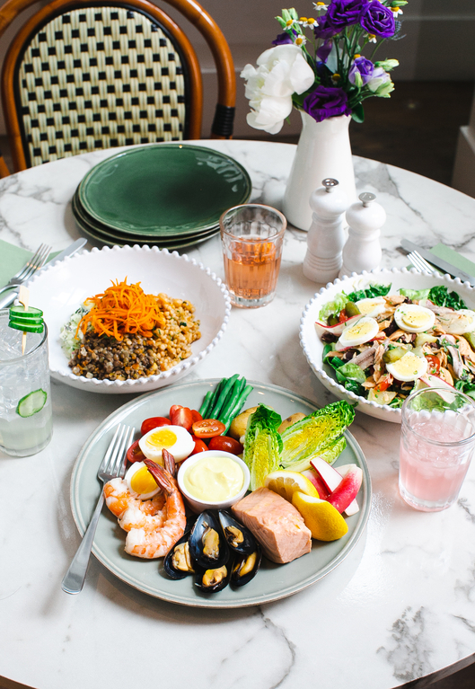 A marble table set with a colorful seafood platter, a quinoa bowl, a salad, glasses of iced tea, and a vase of purple and white flowers.