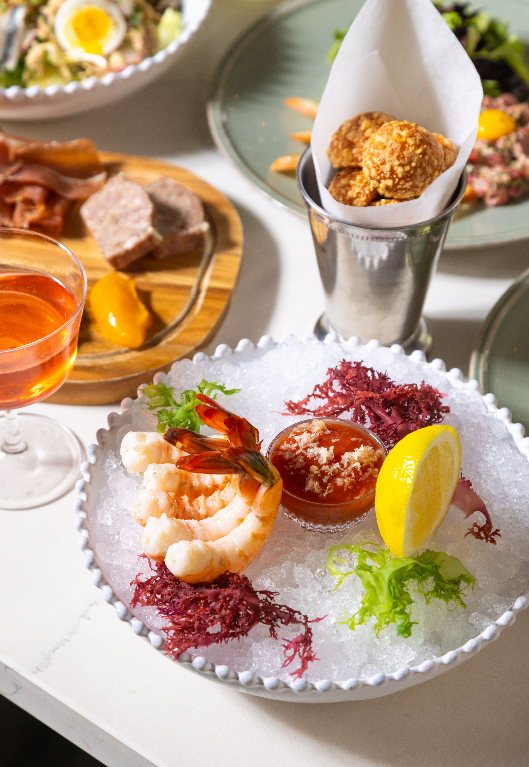 A marble table set with a colorful seafood platter, a quinoa bowl, a salad, glasses of iced tea, and a vase of purple and white flowers.