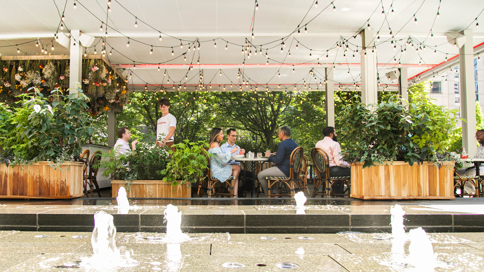 Outdoor patio dining area with string lights, fountain, wooden planters, and guests seated at tables surrounded by greenery