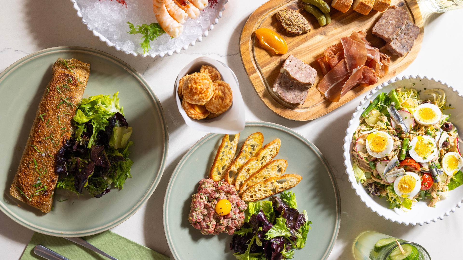 Overhead view of a French bistro-style spread featuring steak tartare with crostini, a crispy crepe with mixed greens, a charcuterie board with cured meats and cornichons, a Niçoise salad with soft-boiled eggs, a shrimp cocktail, and a small bowl of savory biscuits on a white marble table.