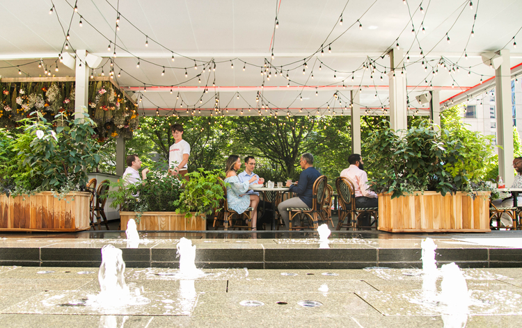 Outdoor patio dining area with string lights, fountain, wooden planters, and guests seated at tables surrounded by greenery