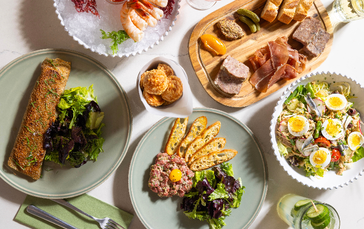 Overhead view of a French bistro-style spread featuring steak tartare with crostini, a crispy crepe with mixed greens, a charcuterie board with cured meats and cornichons, a Niçoise salad with soft-boiled eggs, a shrimp cocktail, and a small bowl of savory biscuits on a white marble table.